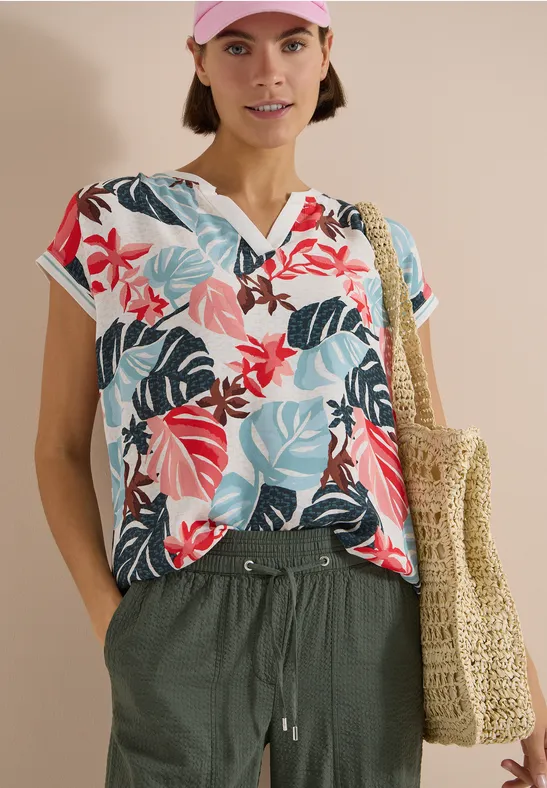 Eine Frau trägt ein buntes Blusen-Shirt mit tropischem Muster, grüne Hose und geflochtene Tasche.