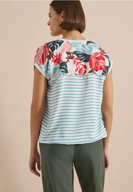 Rückansicht einer Frau mit Kurzarmshirt in blau-weißen Streifen und Blumenmuster, dazu olivgrüne Hose.
