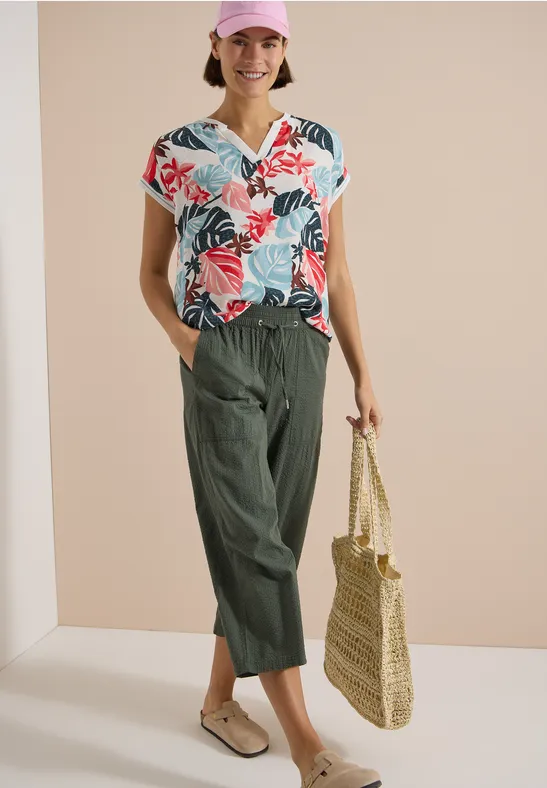 Frau trägt buntes, florales Top, weite grüne Hose und hält eine Strohtasche.