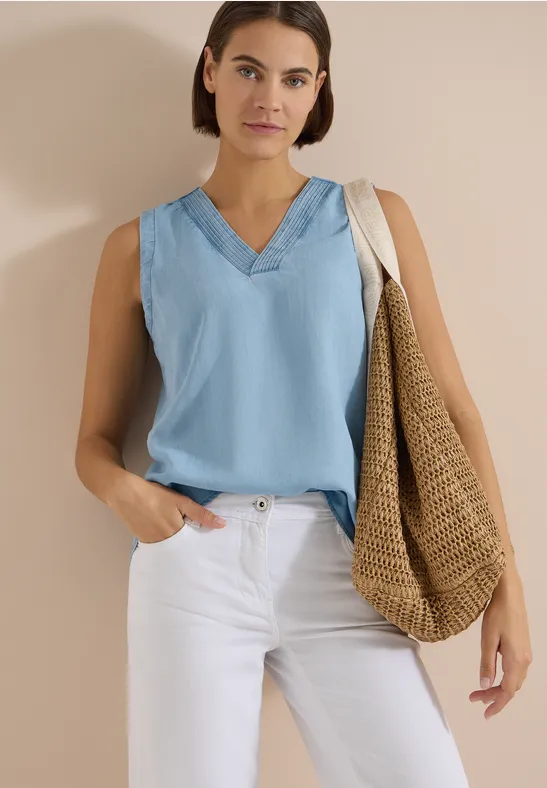 Frau in hellblauem ärmellosen V-Ausschnitt-Top, weißen Hosen und großer Strohtasche vor neutralem Hintergrund.