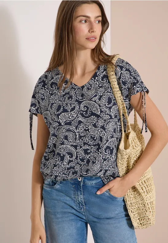 Vrouw in blauwe paisley-blouse met knoopmouwen, spijkerbroek en gevlochten strooitas.