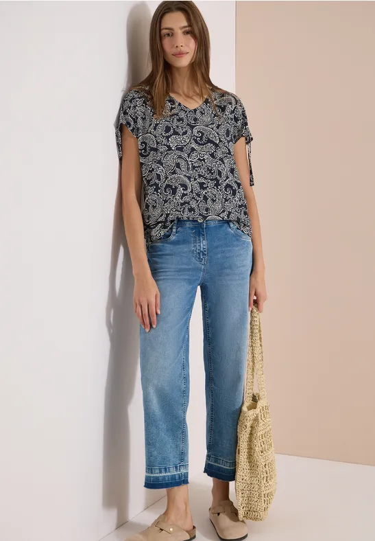 Vrouw in rechte jeans, kort mouwtop met print en gevlochten tas, staand voor een lichte achtergrond.