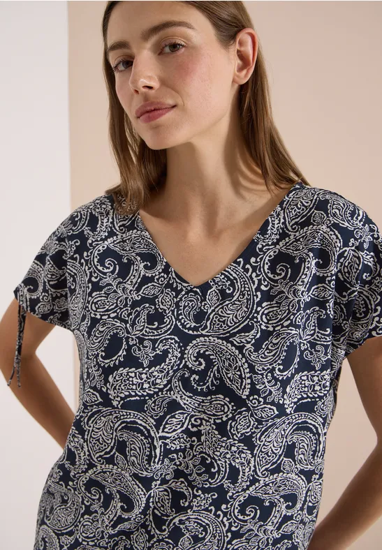 Vrouw draagt donkerblauw overhemdje met paisleyprint en V-hals tegen neutrale achtergrond.