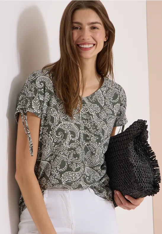 Vrouw in een groen paisleyblouse met korte mouwen, witte broek en een zwarte gehaakte clutch.