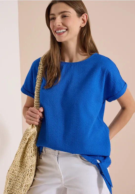 Vrouw in los kobaltblauw shirt met knoopdetail, witte jeans en grote stro tas voor neutrale achtergrond.