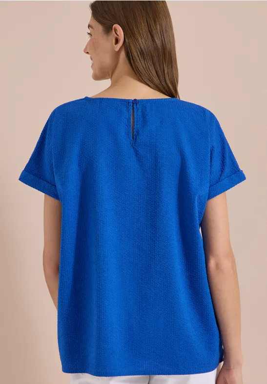 Een vrouw van achteren draagt een los, felblauw shirt met korte mouwen en ronde hals.