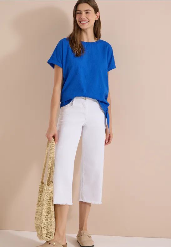 Vrouw voor beige achtergrond met blauwe t-shirt, witte culottes, espadrilles en een gevlochten tas.