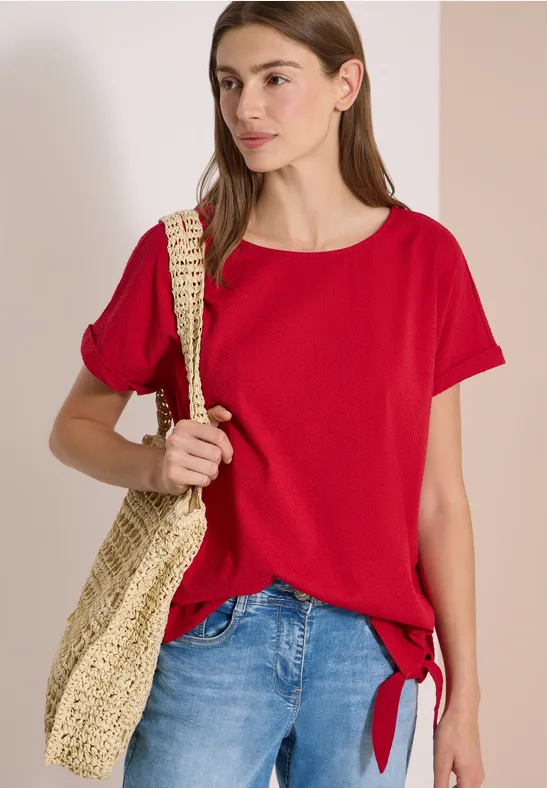 Vrouw in rood shirt met knoop aan de zijkant, blauwe spijkerbroek en gevlochten tas over de schouder.