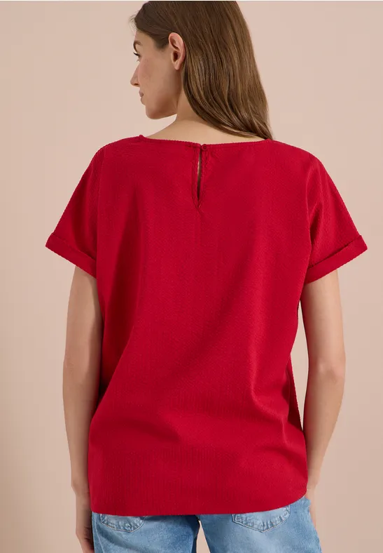 Achteraanzicht van een vrouw in een rood T-shirt met korte mouwen en een blauwe jeans tegen een beige achtergrond.