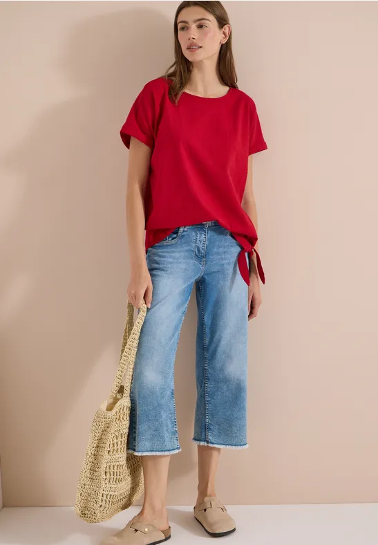 Vrouw in rood T-shirt, wijd korte spijkerbroek, beige instappers en gevlochten tas voor beige achtergrond.