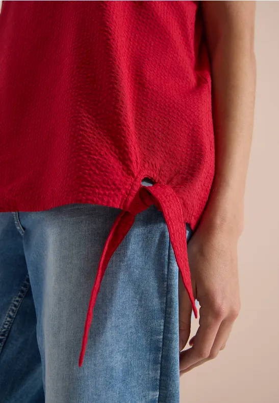 Rode getextureerde mouwloze top met strikdetail onderaan, gecombineerd met een blauwe jeans.