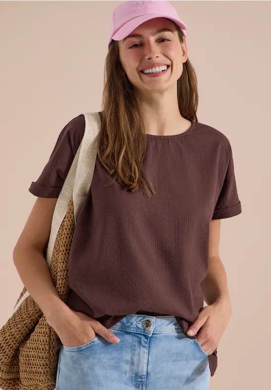 Een jonge vrouw draagt een bruin t-shirt, spijkerbroek en een roze pet, met een strooien tas over haar schouder.