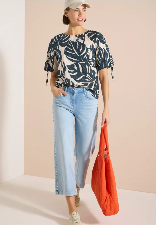 Femme en chemisier à motif de feuilles, jean large clair, baskets vertes et sac rouge.
