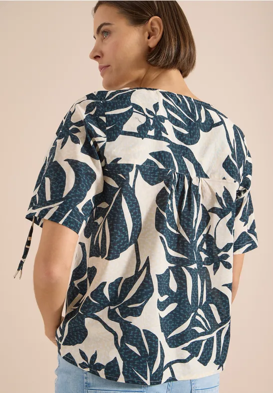 Femme porte un chemisier blanc à motif de feuilles bleu foncé, manches mi-longues et liens sur les côtés.