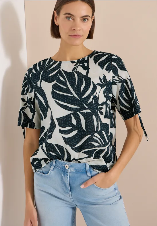 Femme en T-shirt blanc à manches courtes imprimé de feuilles vert foncé, rentré dans un jean bleu clair devant fond beige.
