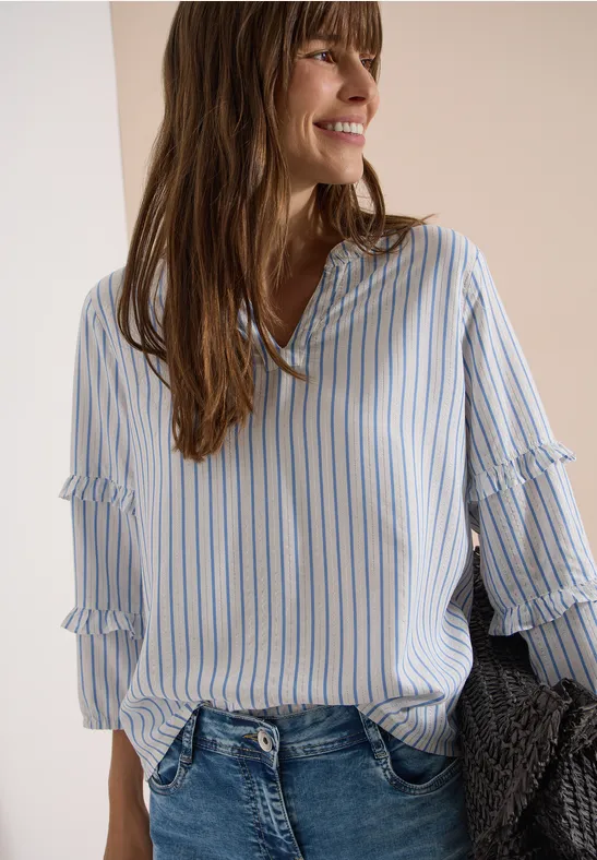 Frau trägt locker sitzende, blau-weiß gestreifte Bluse und Jeans, hält grauen Strickpullover.