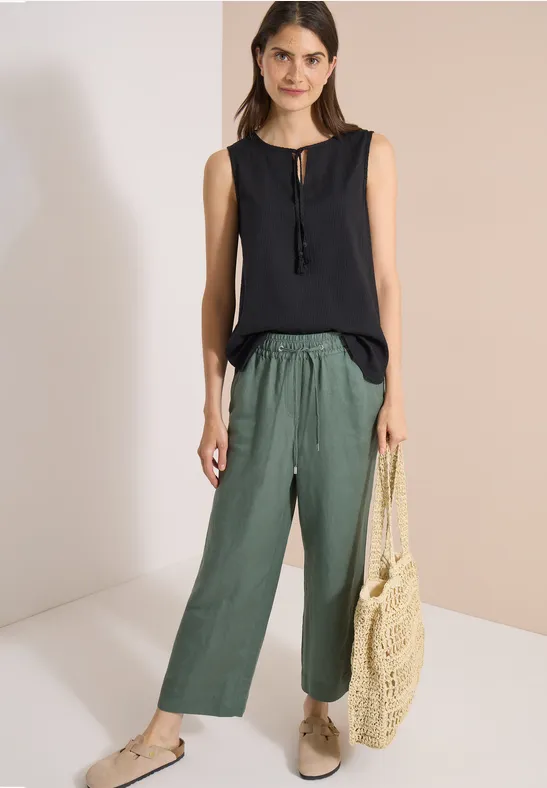Frau trägt schwarzen ärmellosen Top, weite grüne Hose, beige Schuhe und eine Netz-Tasche.