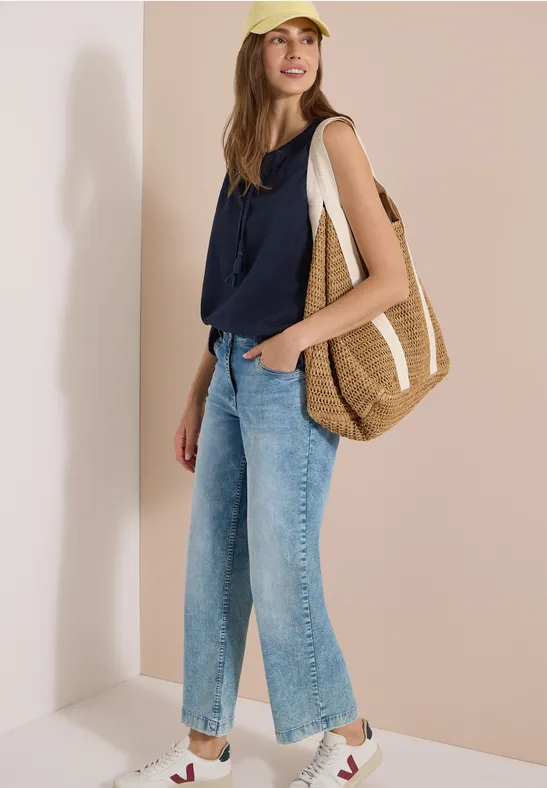 Frau lehnt an einer beigen Wand, trägt dunkelblaues Top, weite Jeans, weiße Sneaker und eine große geflochtene Tasche.