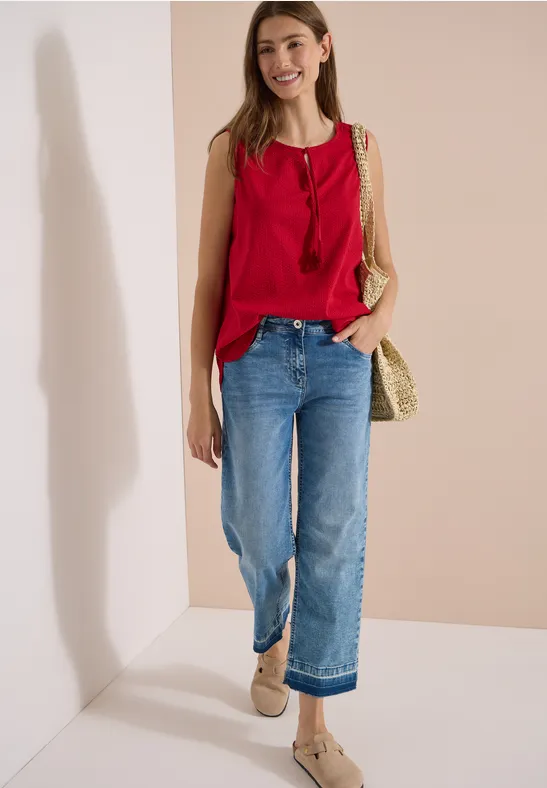 Frau in rotem ärmellosem Top, weiten blauen Jeans und geflochtener Strohtasche vor heller Wand.