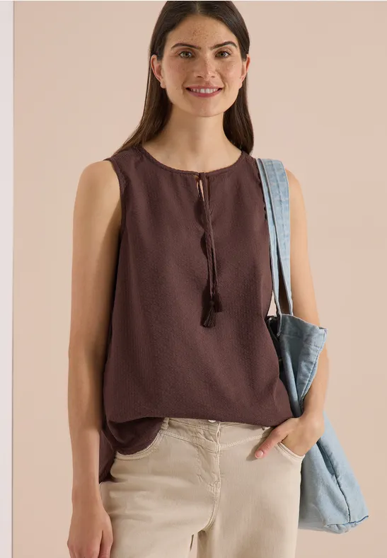 Frau trägt ärmelloses, braunes Top mit Quasten, helle Hose und blaue Jeans-Tasche über der Schulter.