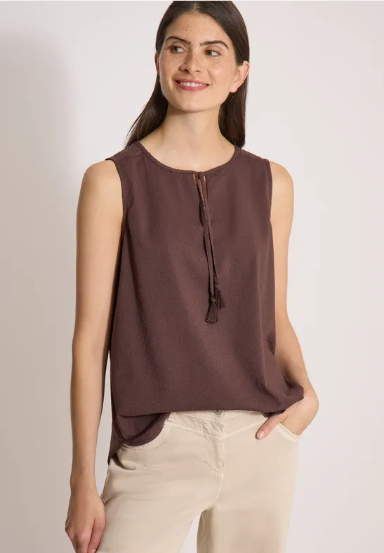 Eine Frau trägt eine ärmellose braune Bluse und beige Hosen und posiert lässig mit einer Hand in der Tasche.