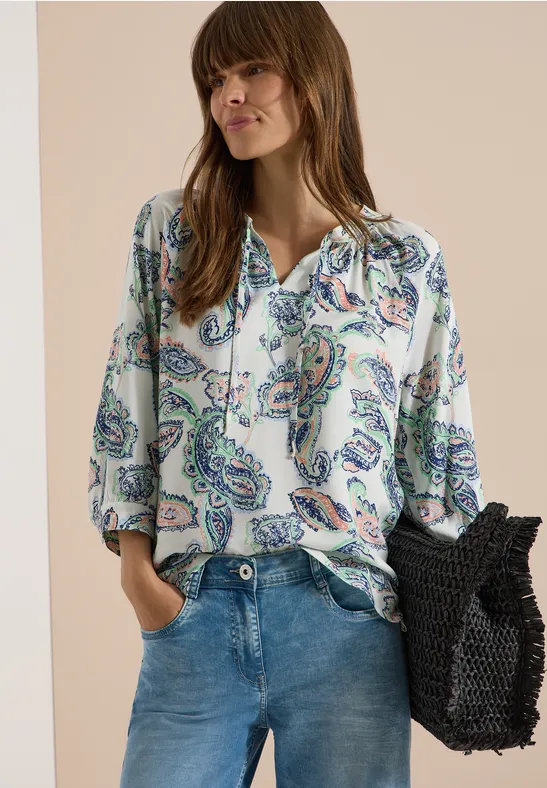 Frau in Jeans und gemusterter Bluse mit Paisley-Print, trägt eine schwarze Netztasche.