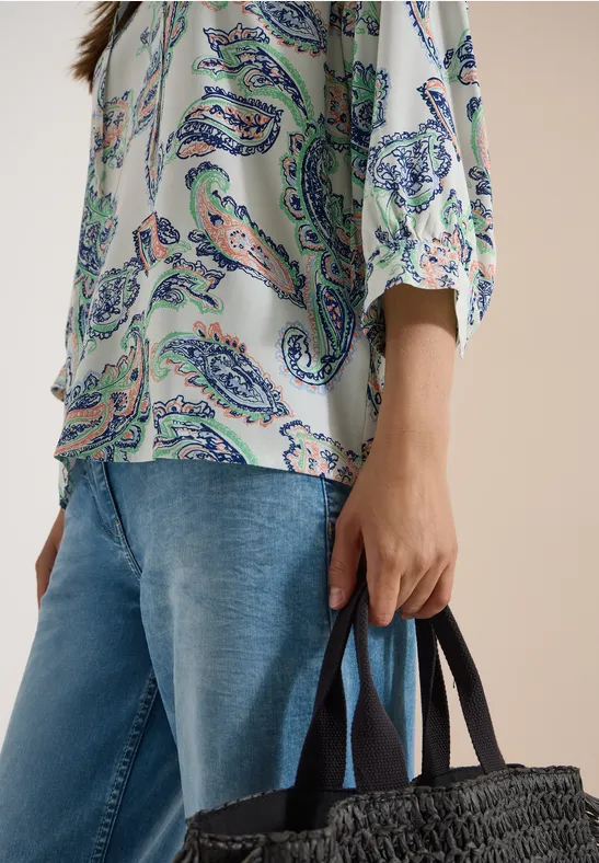 Person in gemusterter Bluse und Jeans hält eine schwarze geflochtene Korbtasche.