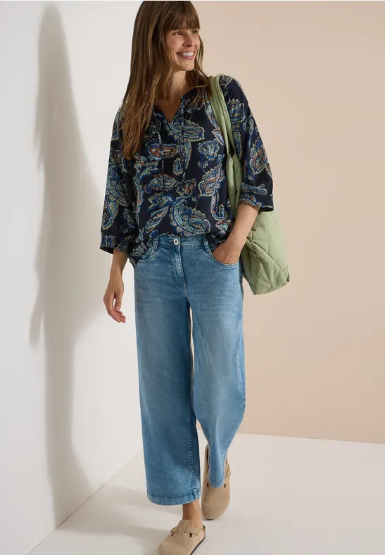 Frau in gemusterter Bluse, weitem Jeans, Stiefeletten und grüner Tasche vor neutralem Hintergrund.