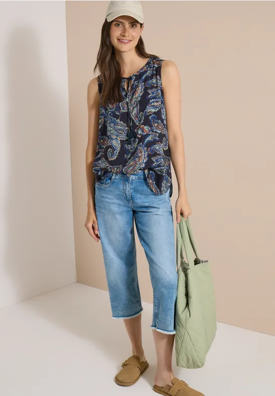 Eine Frau trägt ein ärmelloses, blau-weiß gemustertes Top, Capri-Jeans, bequeme Sandalen und eine grüne Tasche.