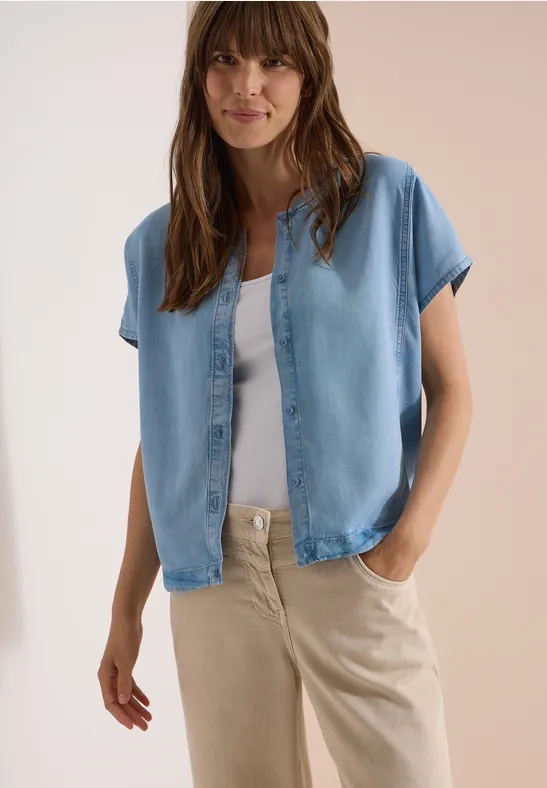 Vrouw in lichtblauw kort mouwen overhemd op een wit topje en beige broek, ontspannen pose.