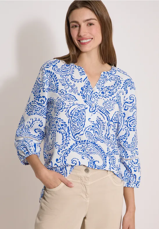 Vrouw in witte blouse met blauw paisley-patroon en beige broek tegen neutrale achtergrond.