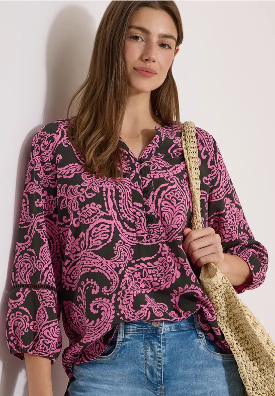 Vrouw met roze paisley-blouse, spijkerbroek en rieten tas.