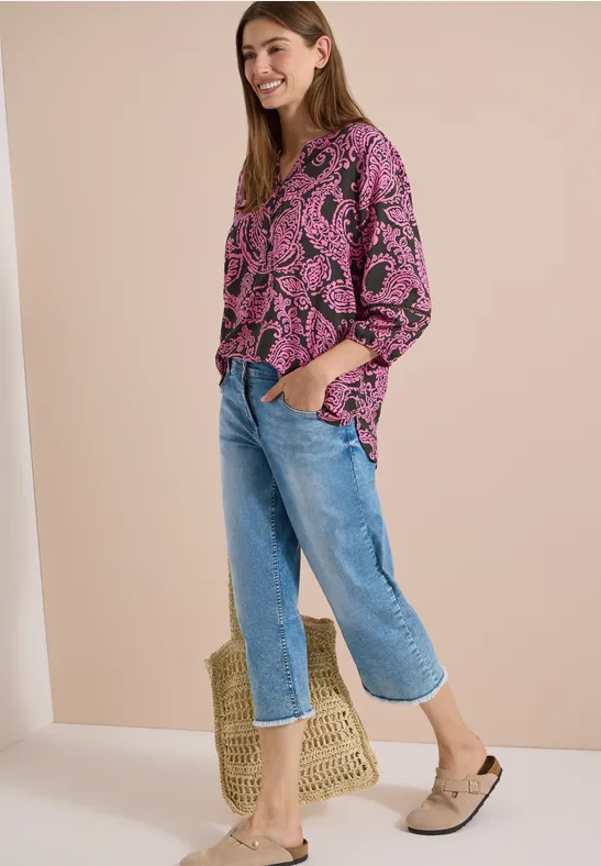 Een vrouw in een roze top met bladprint, blauwe jeans, beige mules en een strooien tas.