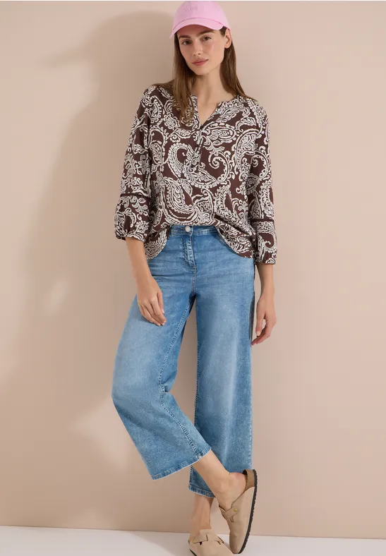 Vrouw in paisleyblouse, wijde blauwe jeans en lichte espadrilles voor een egale achtergrond.