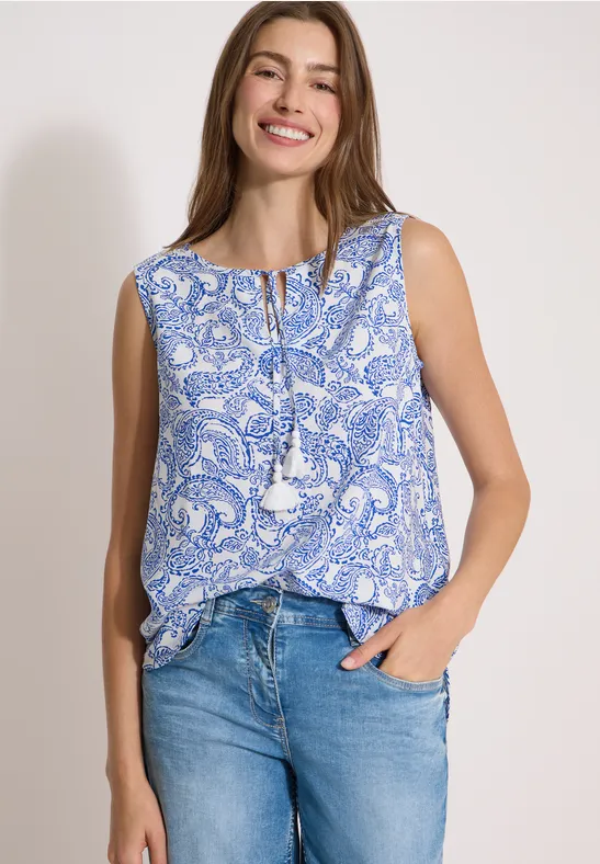 Frau in blau-weißem Paisley-Top mit Quasten und Jeans, vor neutralem Hintergrund.