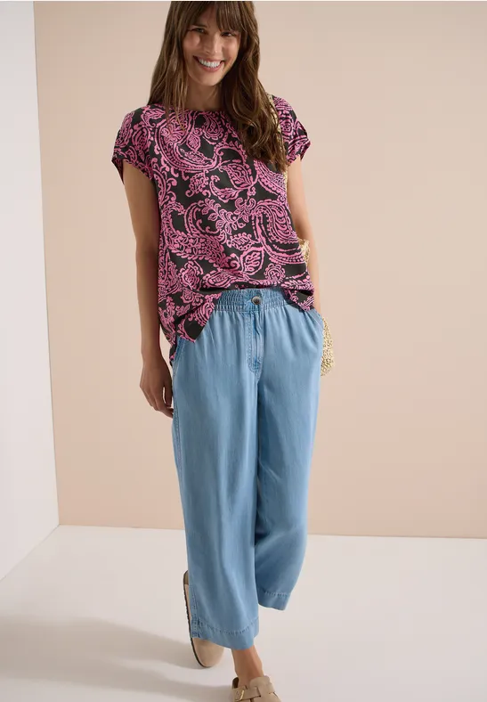 Frau in pink-schwarzem Printshirt und hellblauen, weiten Hose, vor neutralem Hintergrund.