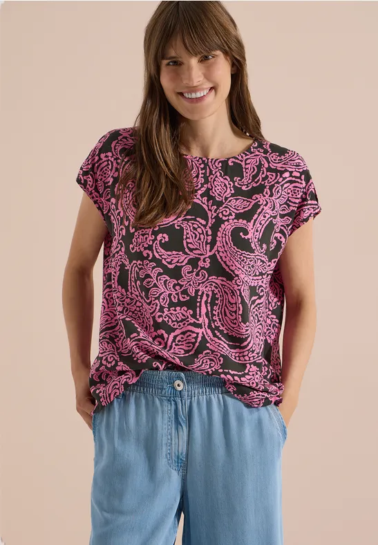 Frau trägt pink gemustertes T-Shirt und hellblaue Jeanshose, steht mit Händen in den Taschen.