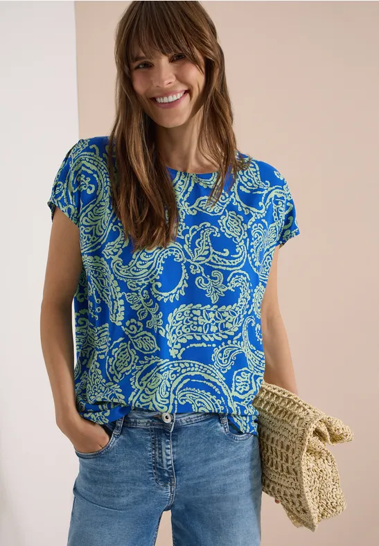 Frau trägt blaues, gemustertes T-Shirt, Jeans und hält eine geflochtene Strohtasche.