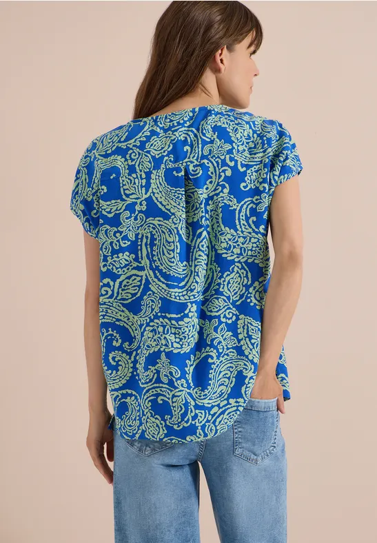 Frau von hinten in blauer Kurzarmbluse mit gelbgrünem Paisley-Muster und Jeans.