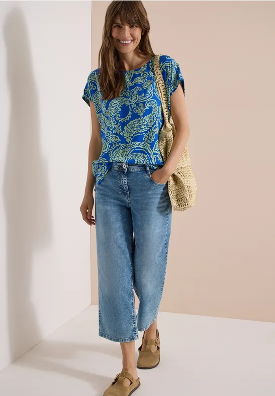 Frau trägt blau gemustertes T-Shirt, helle Culotte-Jeans, Strohtasche und beige Keilsandalen.
