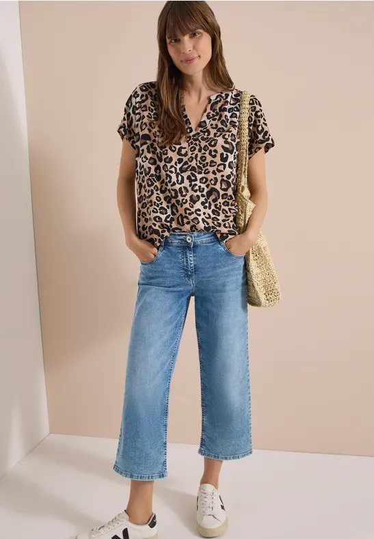 Frau mit Leopardenbluse, weiten Jeans-Culottes, weißen Sneakern und Strohtasche vor beiger Wand.