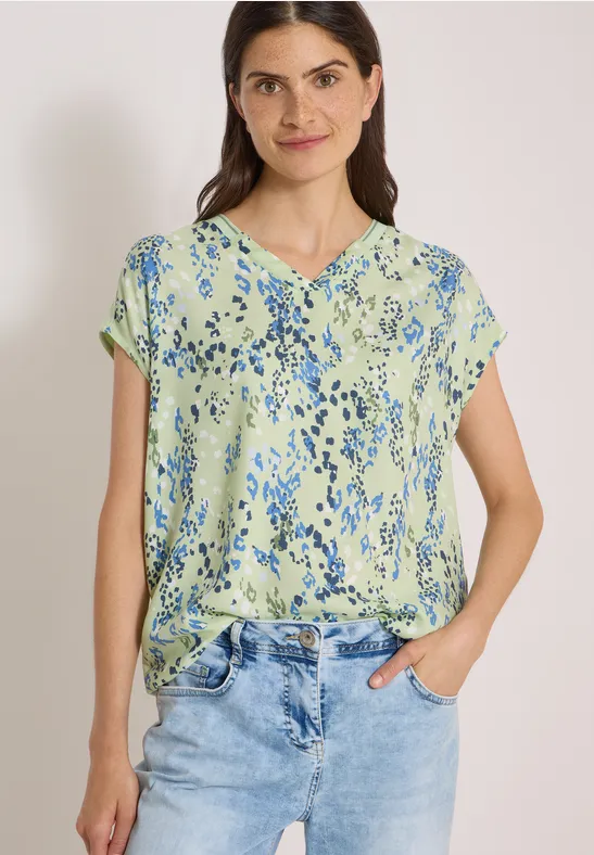 Frau in hellgrüner Bluse mit blauem Blumenmuster und Jeans, lächelnd, Hände in den Taschen.