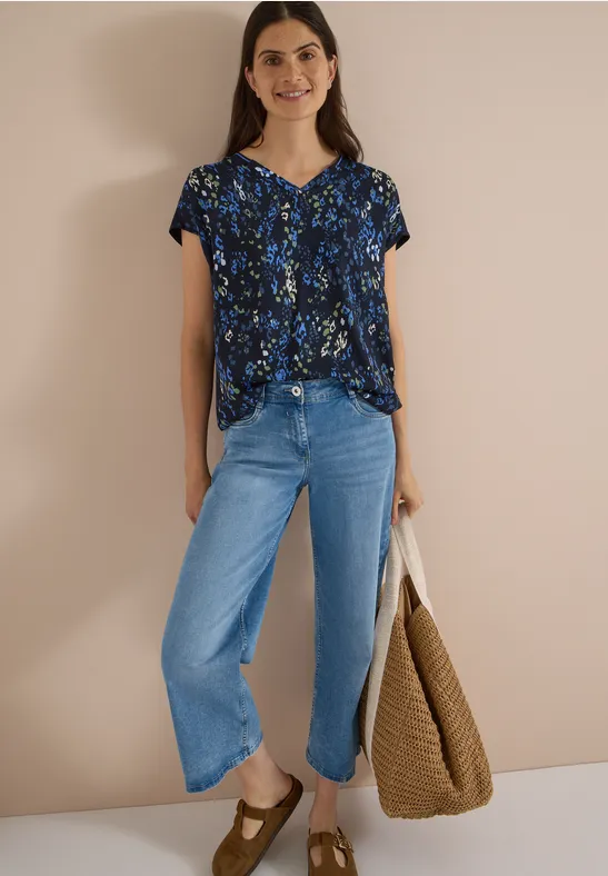 Femme portant un chemisier fleuri, un jean large et un grand sac tressé marron sur fond neutre.