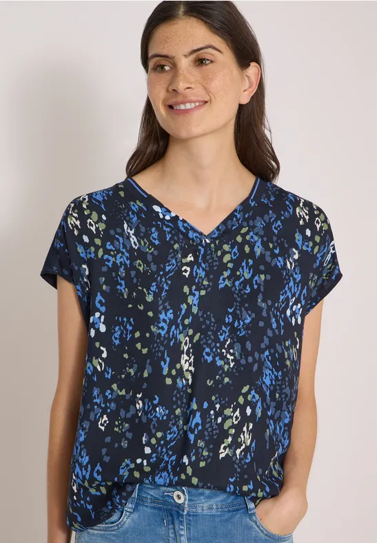 Femme souriante en chemisier noir à motif floral bleu, col V, manches courtes, mains dans les poches.
