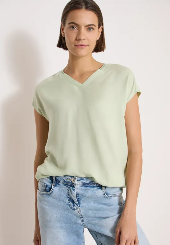 Frau in locker sitzender, hellgrüner V-Neck-Bluse und hellblauen Jeans vor weißem Hintergrund.