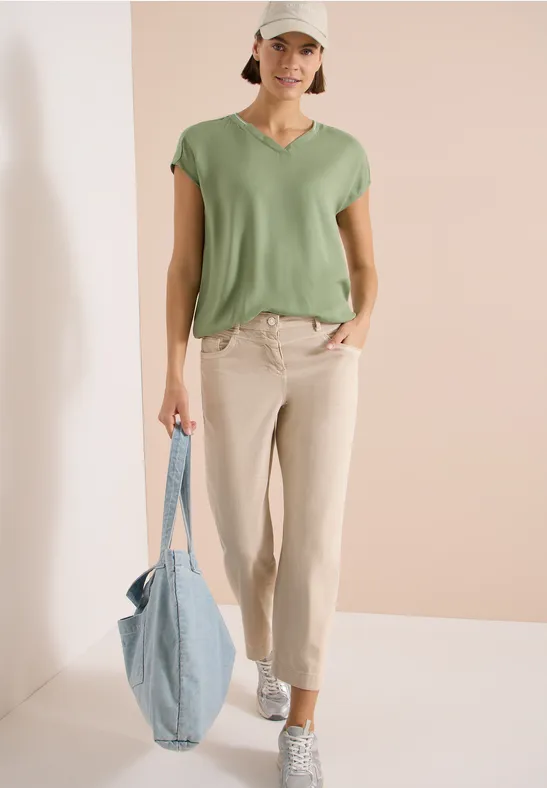Eine Frau trägt ein grünes T-Shirt, helle Hose, weiße Sneaker und hält eine blaue Tragetasche vor schlichtem Hintergrund.