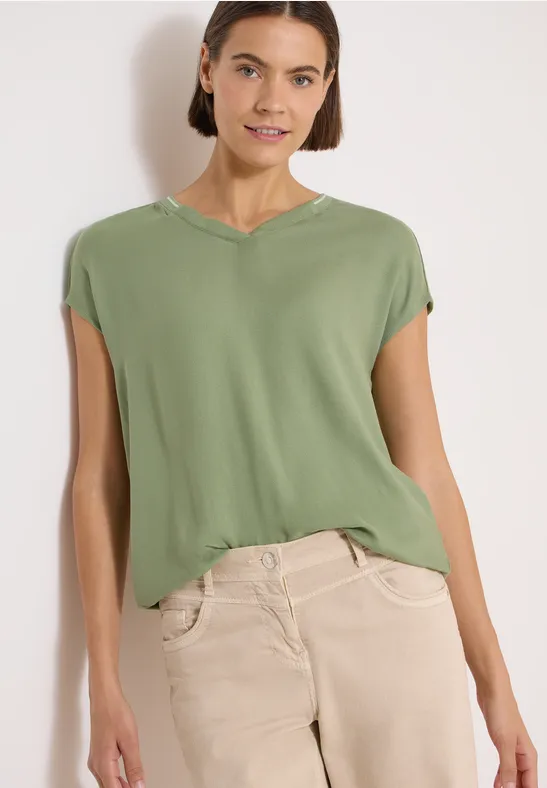 Frau trägt lockeres grünes T-Shirt mit Cap-Ärmeln und beigefarbener Hose.