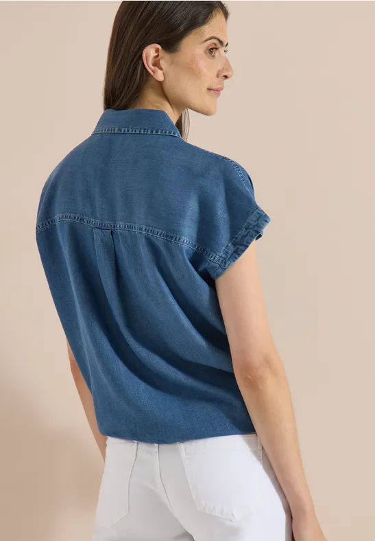 Une femme porte un chemisier en jean à manches courtes et un pantalon blanc, vue de dos sur fond clair.