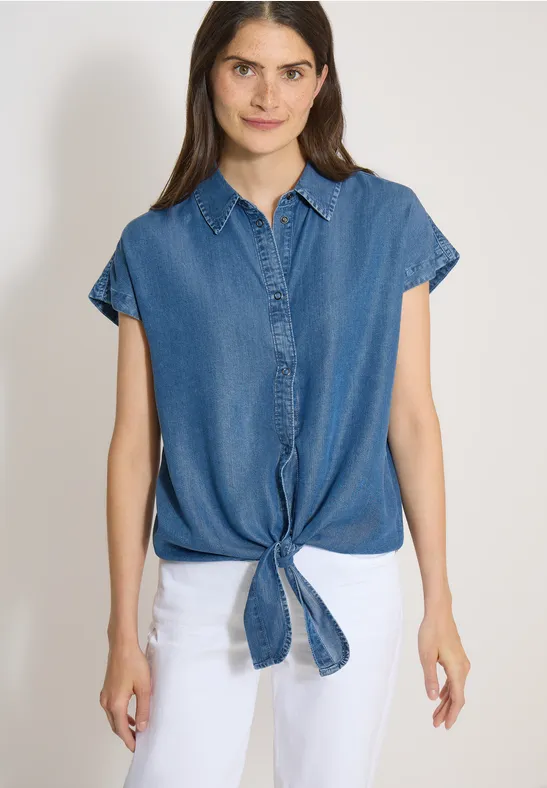 Haut en jean sans manches noué à l'avant, porté avec un pantalon blanc sur fond neutre.