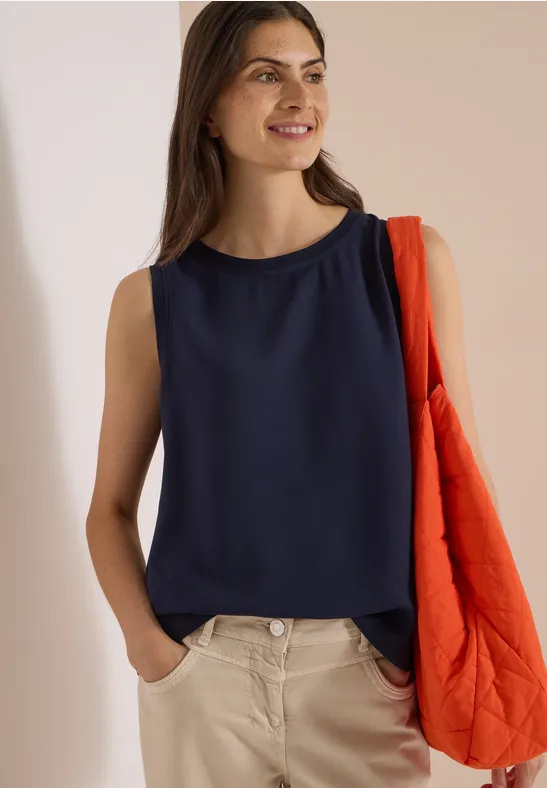 Frau in marineblauem ärmellosem Top und beigen Hosen, lächelnd mit orangener Stepp-Tasche über der Schulter.